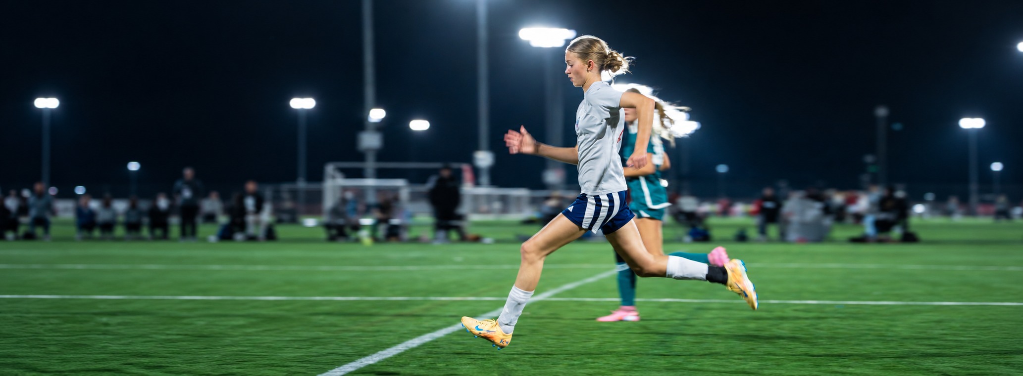 Soccer player in a gray jersey strides across the field under the lights during a match.