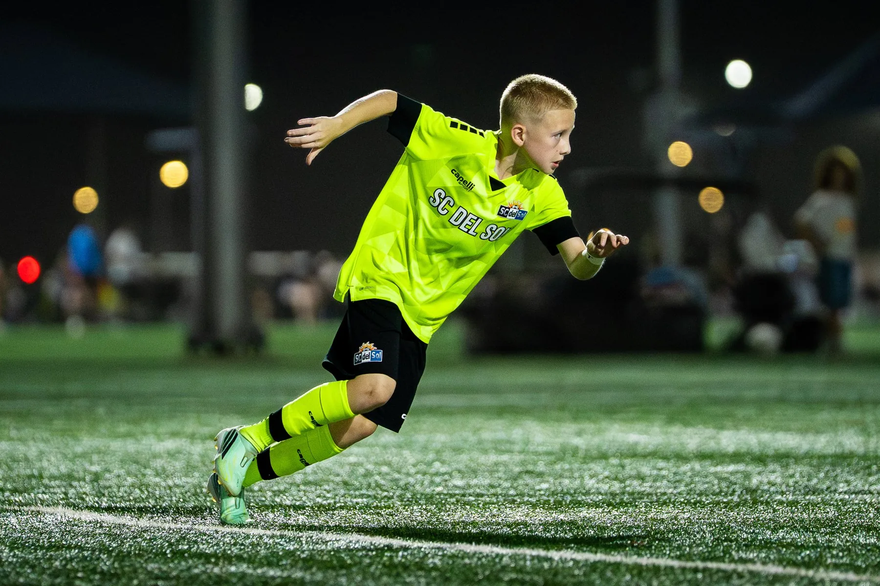 Jamo in his neon yellow soccer uniform runs across illuminated turf during a night game.