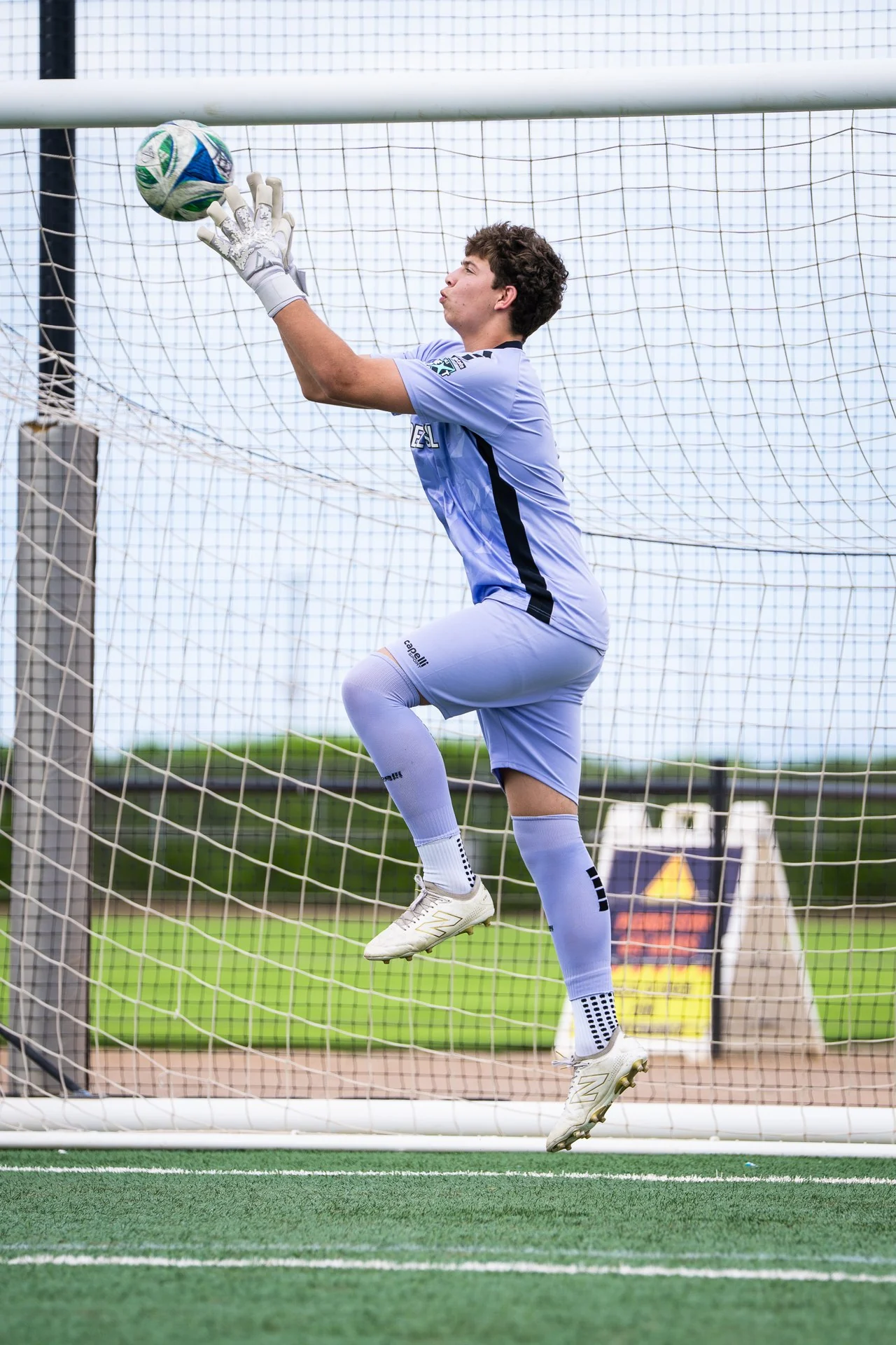 Goalkeeper Stanley jumps with raised gloves to catch the ball, his extended foot and the goal frame visible in daylight.