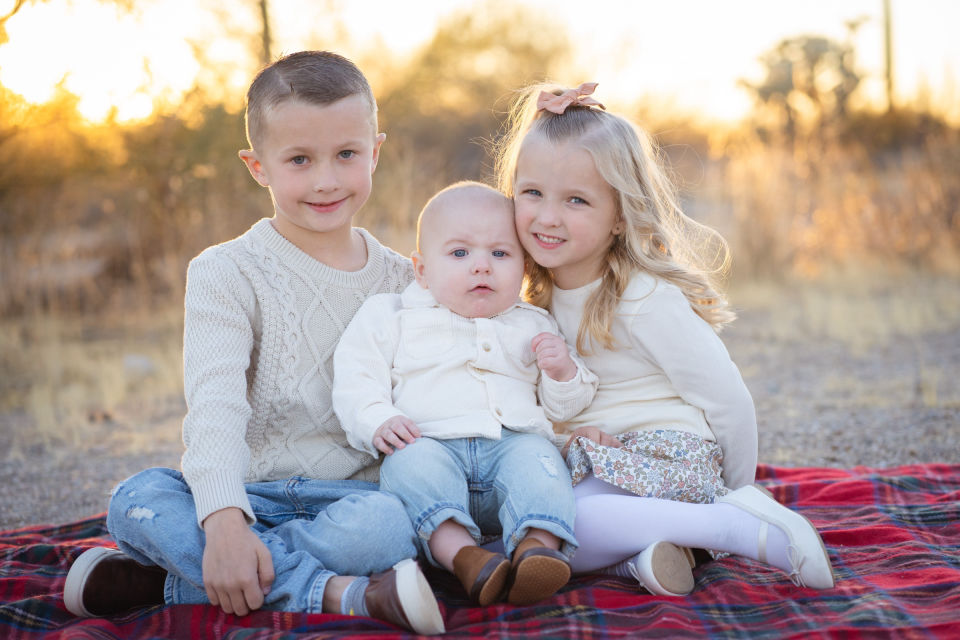 Three siblings sitting together on a blanket in warm evening light.