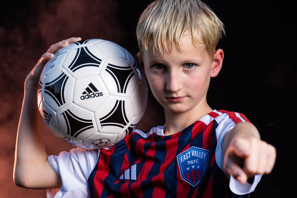 Soccer player points toward the camera with a ball at his shoulder.