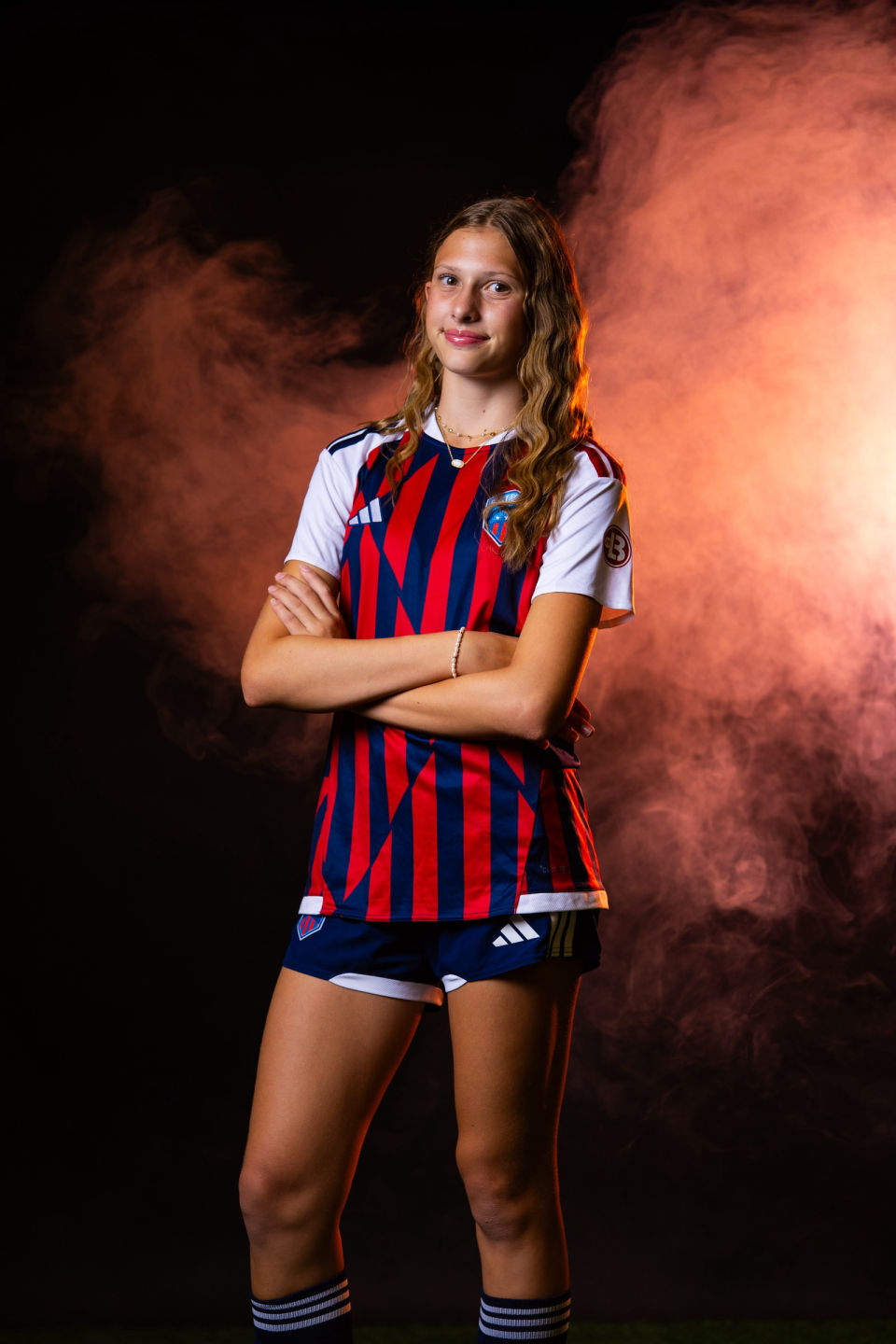 Smiling soccer player poses with folded arms against warm orange smoke.