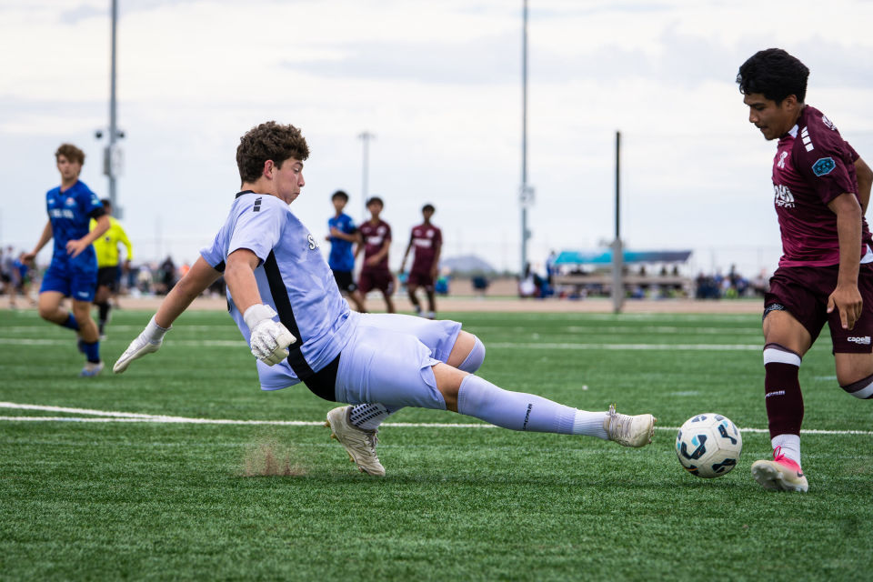 Goalkeeper slides low with an outstretched leg to block an approaching attacker.