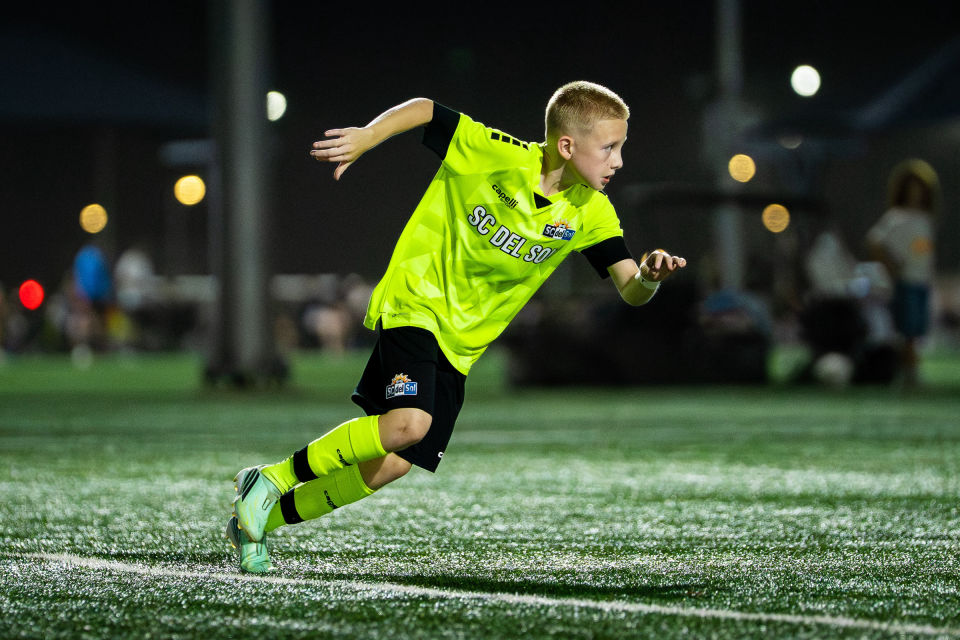 Young soccer player in a bright yellow jersey accelerates across the floodlit field.