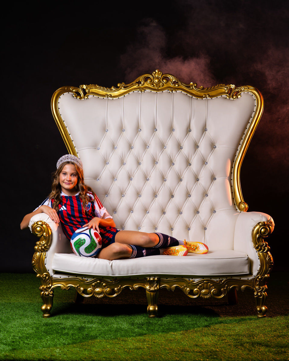 Soccer player seated sideways on a white-and-gold throne with a soccer ball.