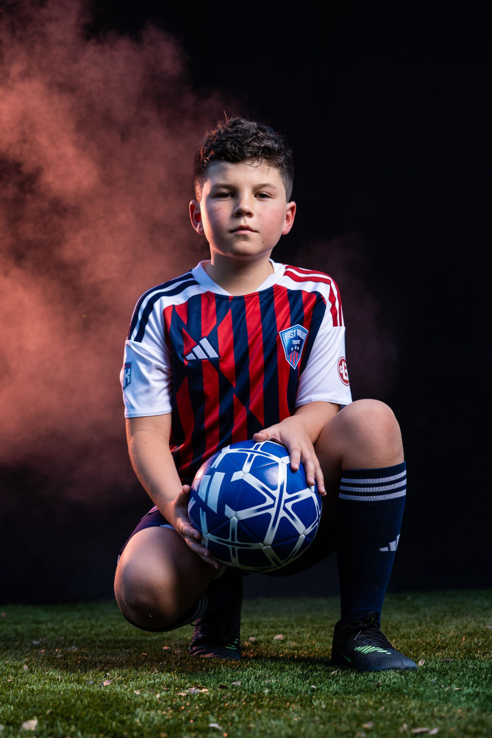 Soccer player kneels with a blue ball against purple and orange smoke.