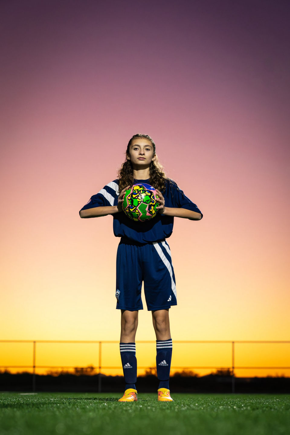 Soccer player holds a ball at chest height against a purple-and-gold sunset.