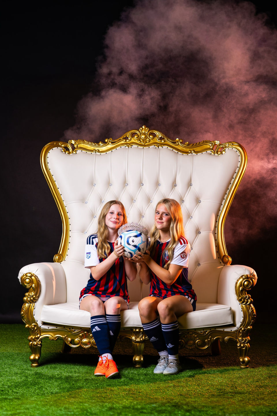 Two smiling soccer teammates hold a crowned soccer ball on a white and gold throne.