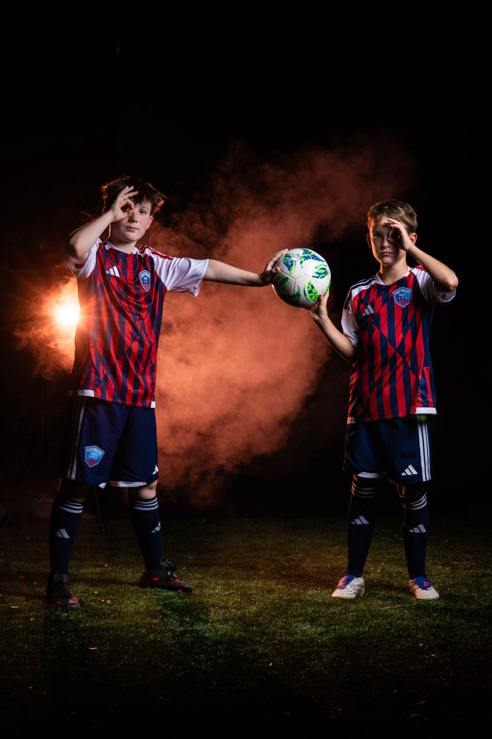 Two soccer teammates salute playfully against orange smoke.