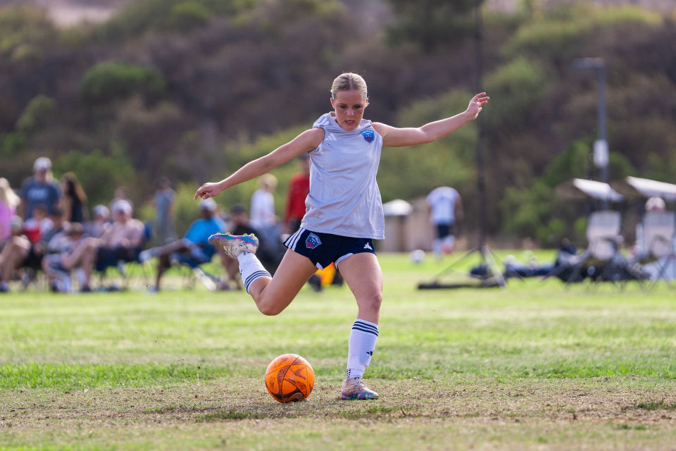 Soccer player in white winds up to strike an orange ball during a daytime match.