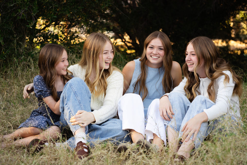 Four sisters laugh together while seated in the grass.
