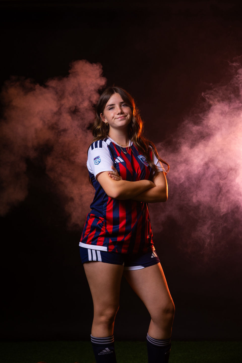 Soccer player poses confidently with folded arms against pink smoke.