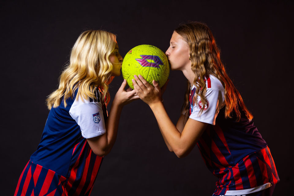 Two teammates kiss a bright yellow soccer ball against a dark backdrop.