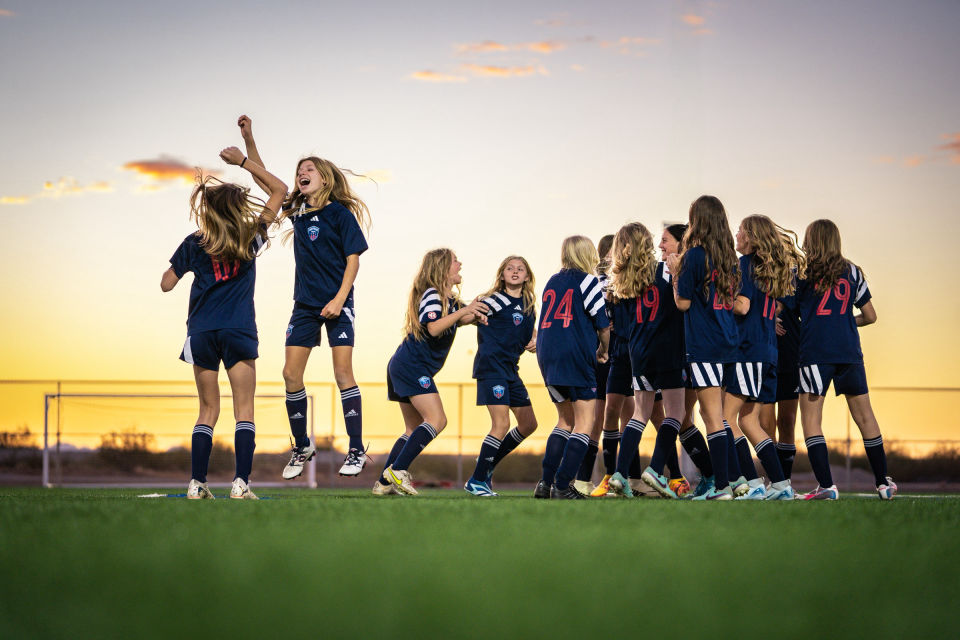 Soccer teammates celebrate with raised hands on the turf beneath evening clouds.