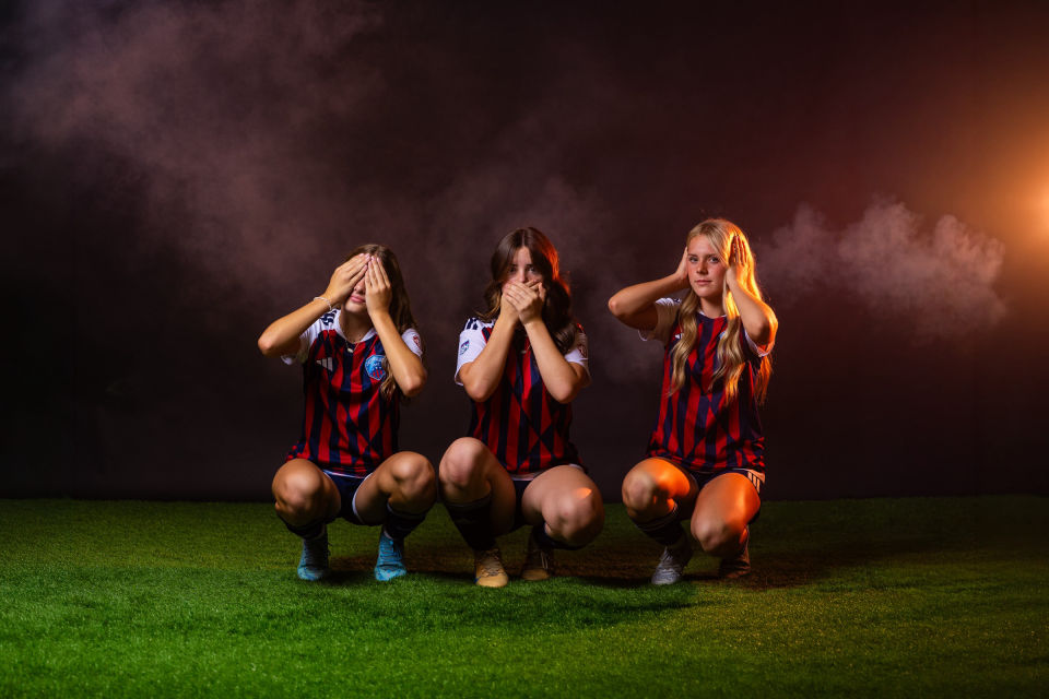 Three soccer teammates pose with eyes, mouth, and ears covered against a smoky backdrop.