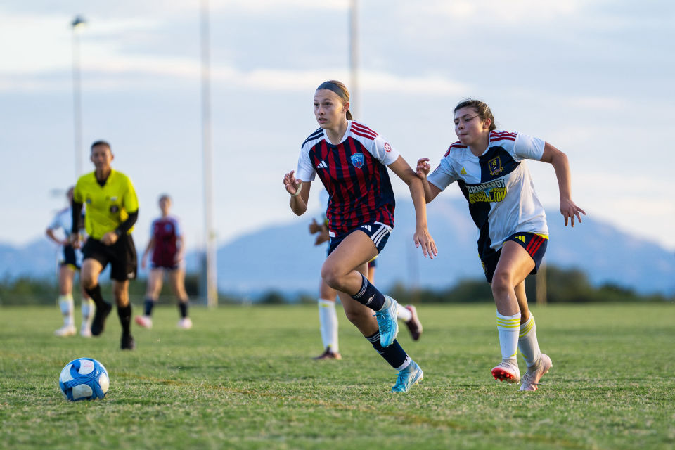 Two soccer players sprint shoulder to shoulder after a blue and white ball.