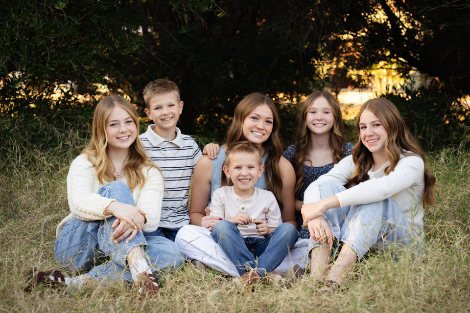 Six siblings sit together in the grass beneath trees.