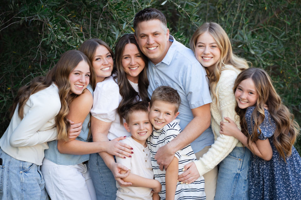 Family shares a close embrace surrounded by green trees.