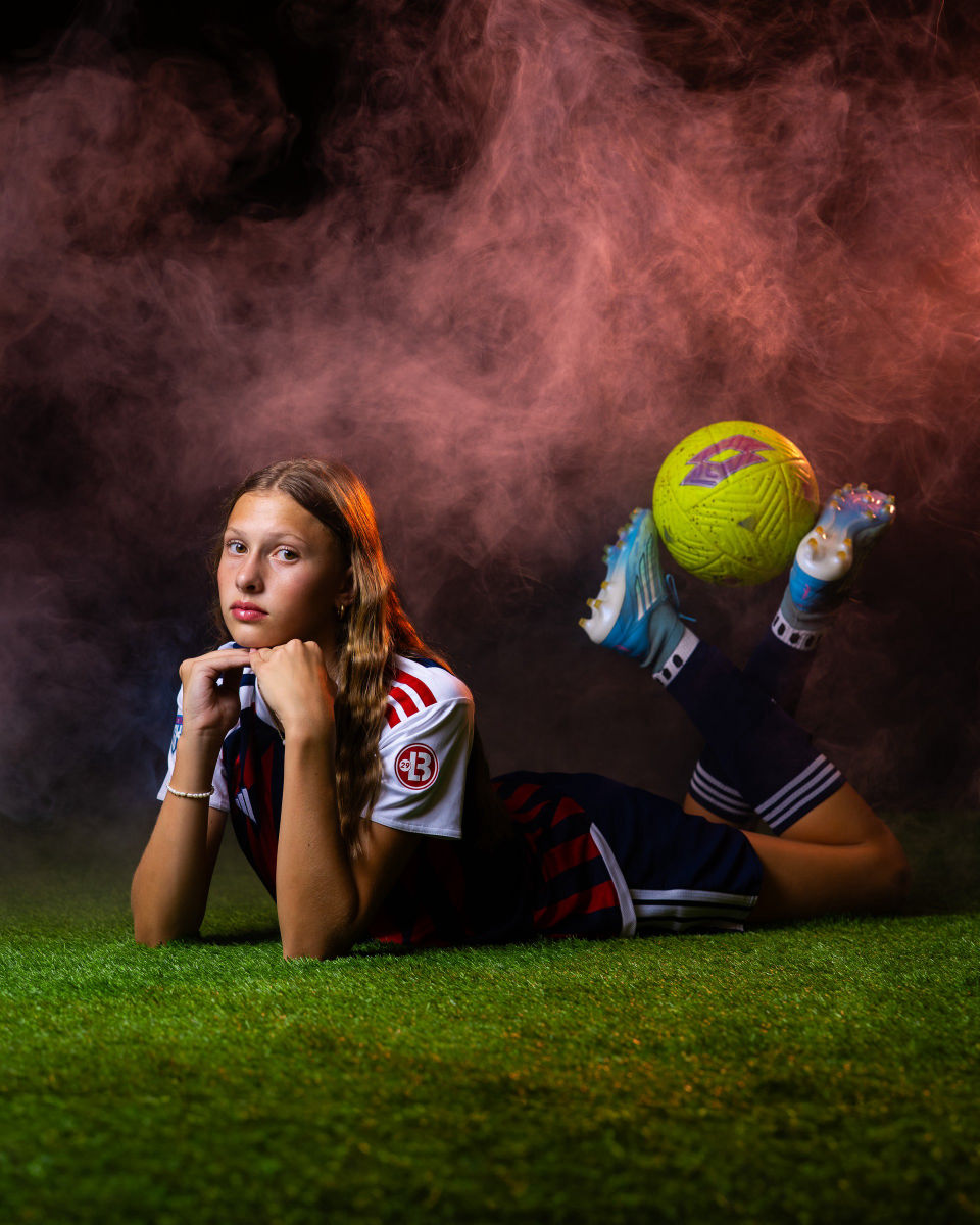 Soccer player resting her chin on her hands with a yellow ball balanced between raised cleats and pink smoke behind her.