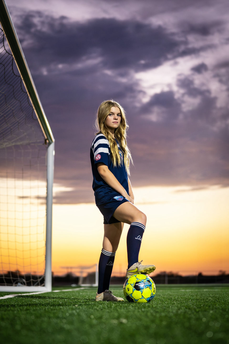Soccer player poses beside the goal with her foot on a bright ball beneath dramatic purple clouds and a golden sunset.