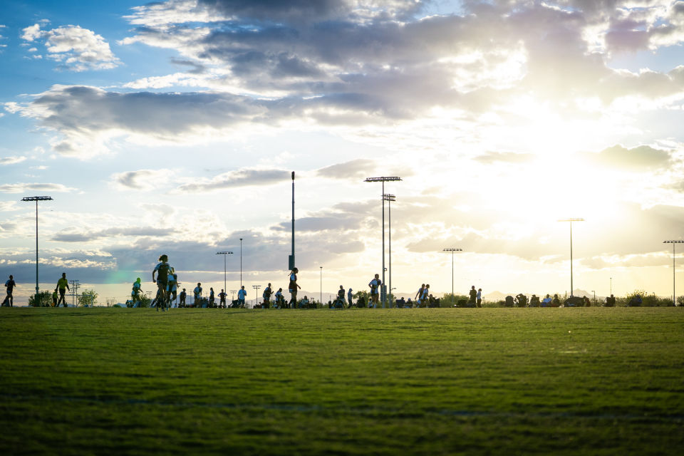 Sunlight breaks through dramatic clouds above soccer fields, silhouetted players, and field lights.