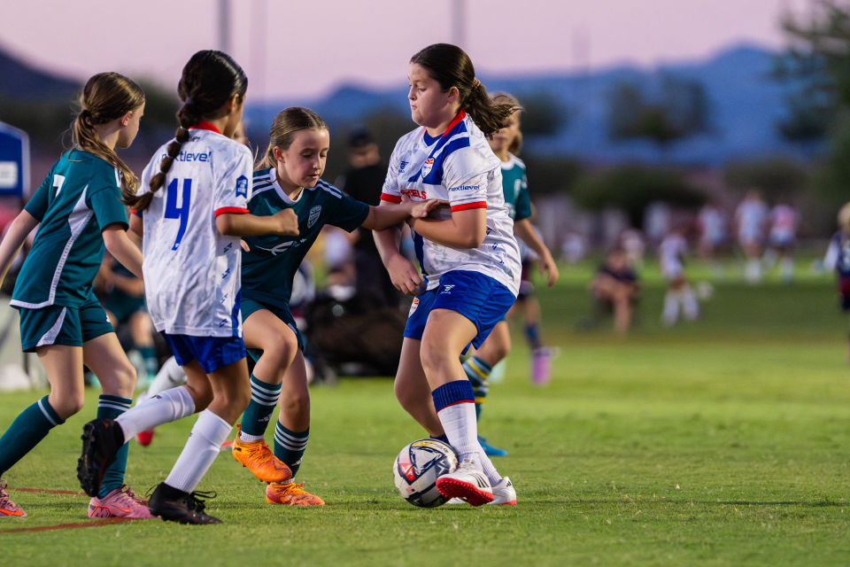 Young soccer player shields the ball from an opponent in evening light.