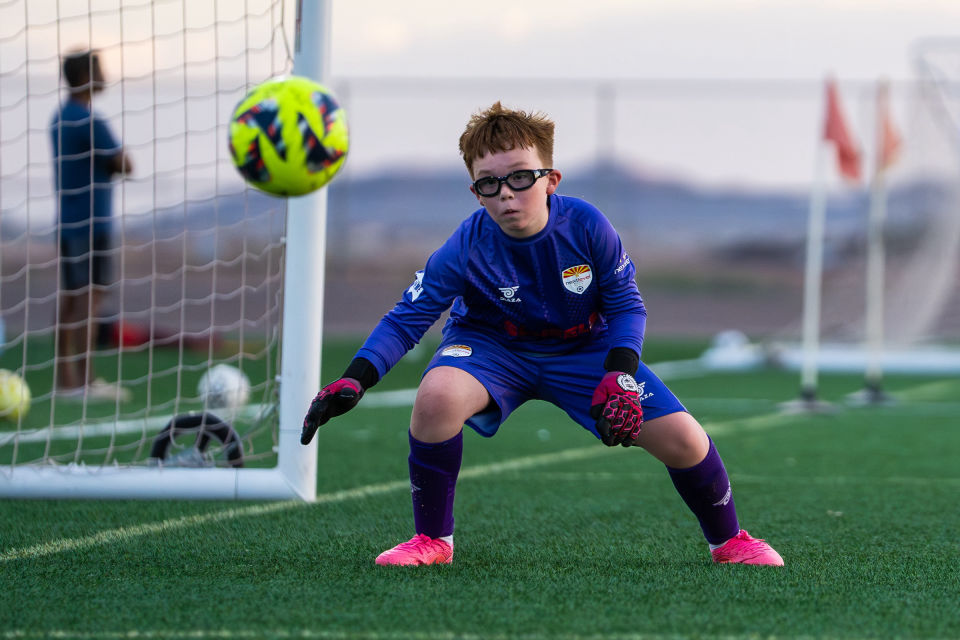 Young goalkeeper crouches with gloved hands ready as the ball approaches.