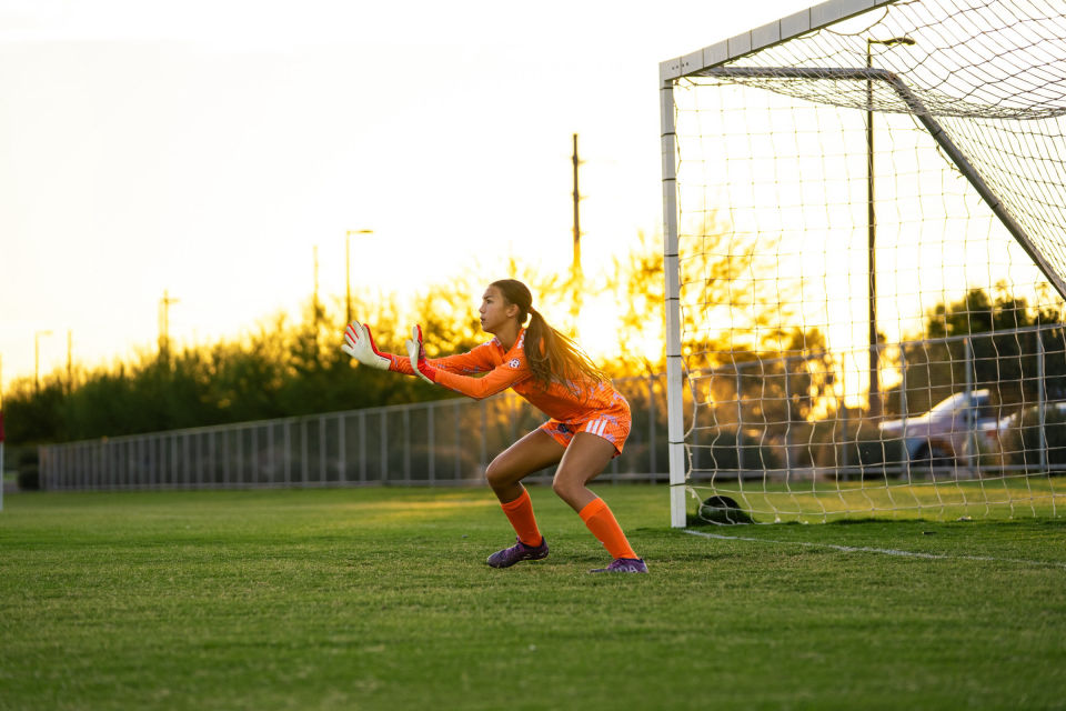 Goalkeeper in orange holds her gloves ready near the goal in golden evening light.
