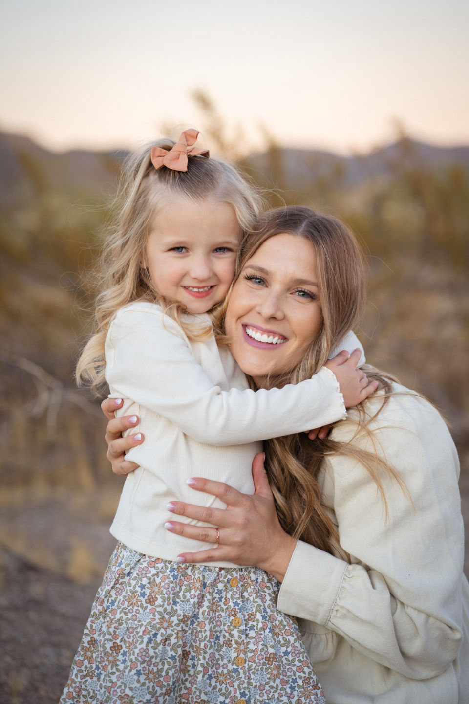 Mother hugs her smiling daughter in warm evening light.