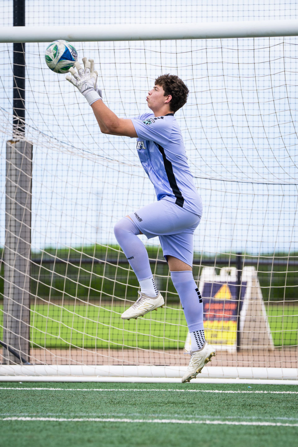 Goalkeeper jumps with both gloves raised to catch the ball near the crossbar.