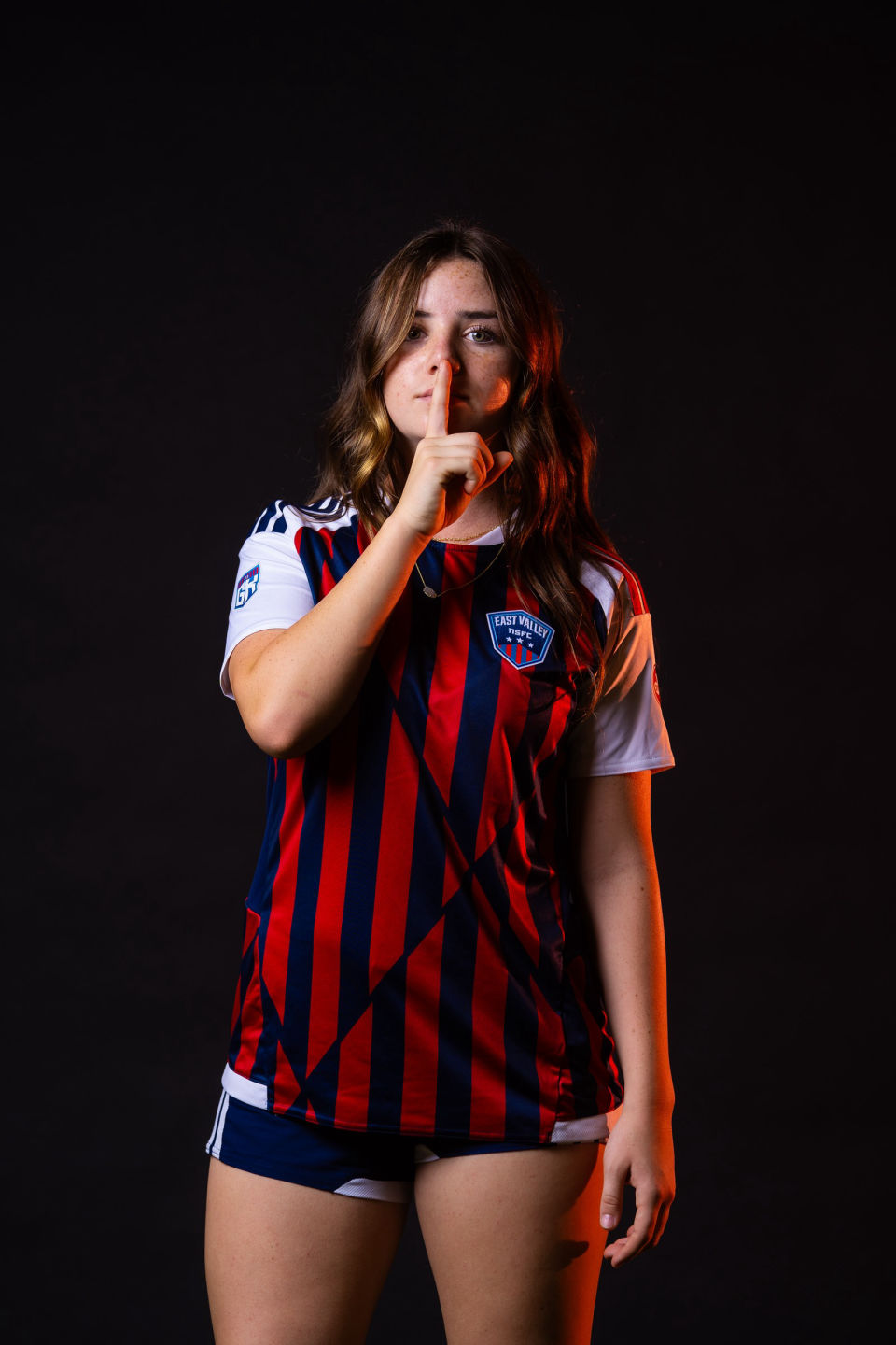 Soccer player holding one finger to her lips, lit with a warm orange rim light.