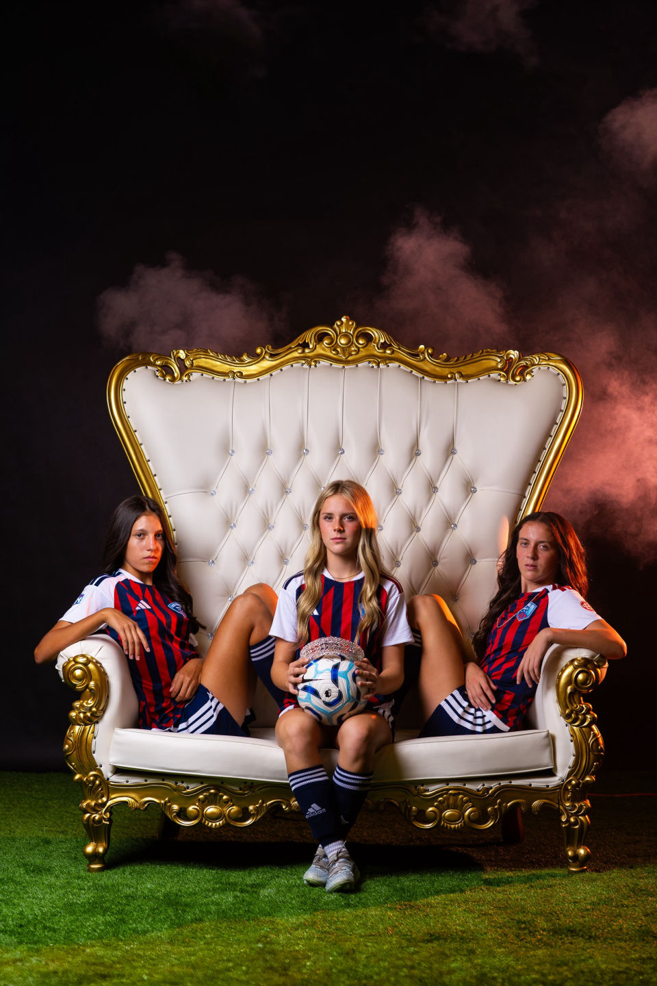 Three soccer teammates sit on a gold-trimmed throne with a crowned ball and pink smoke behind them.
