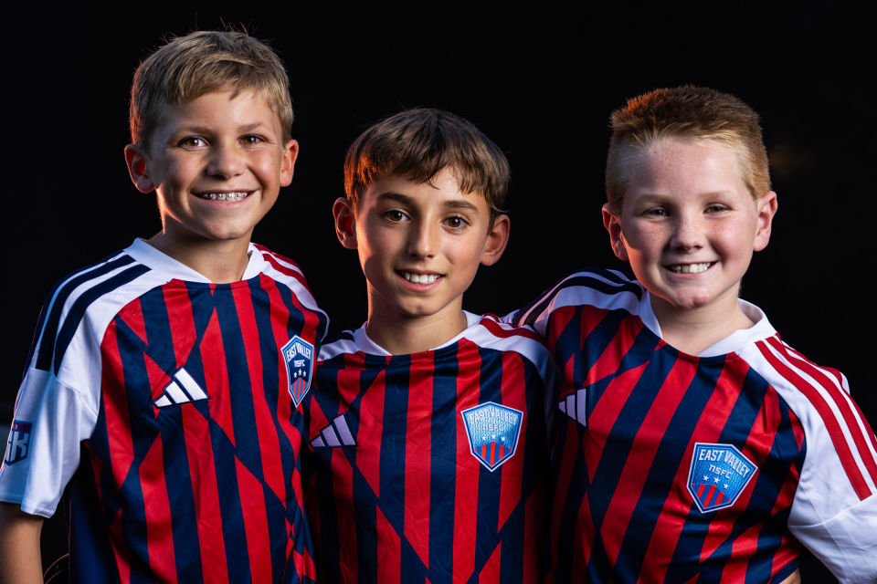 Three smiling soccer teammates stand shoulder to shoulder.