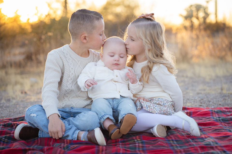 Two siblings kiss their baby sibling while sitting together on a blanket.