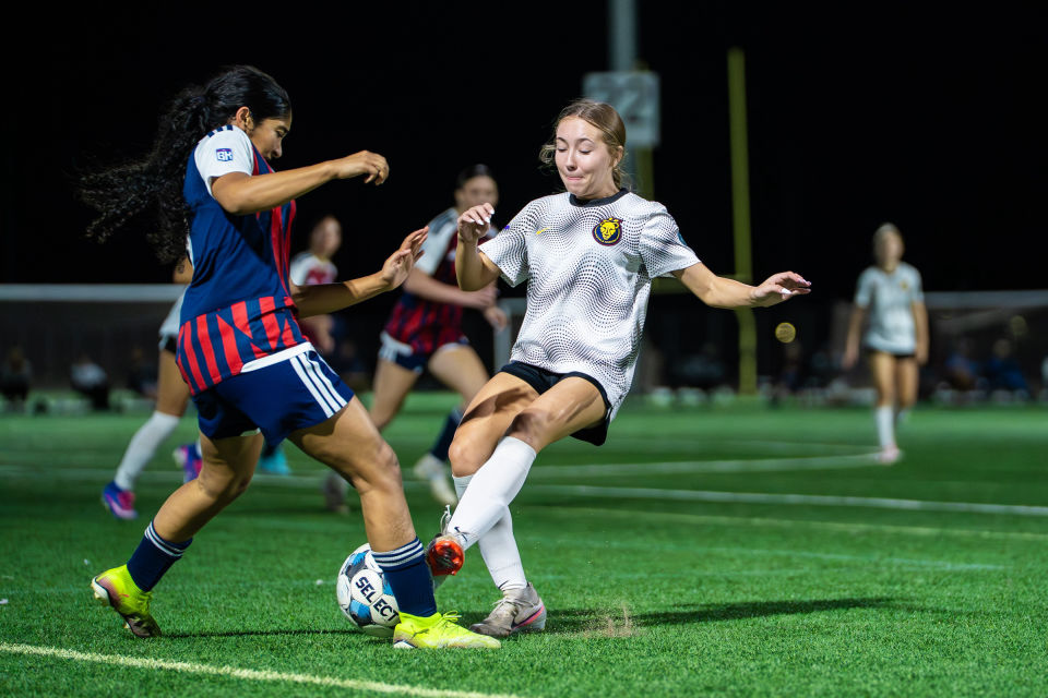 Soccer players in gray and striped jerseys battle for possession under stadium lights.