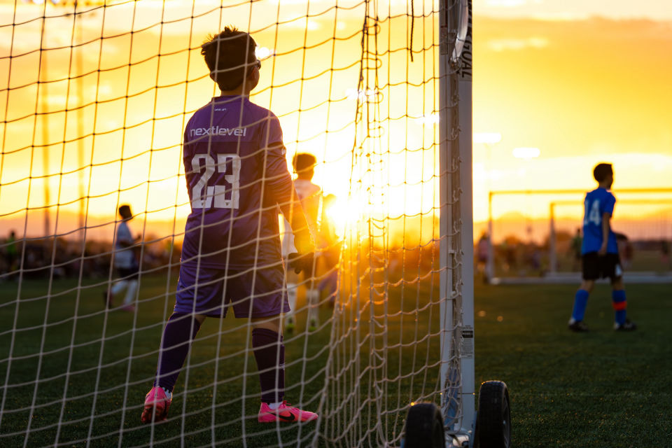 Goalkeeper watches play through the goal net in golden sunset light.